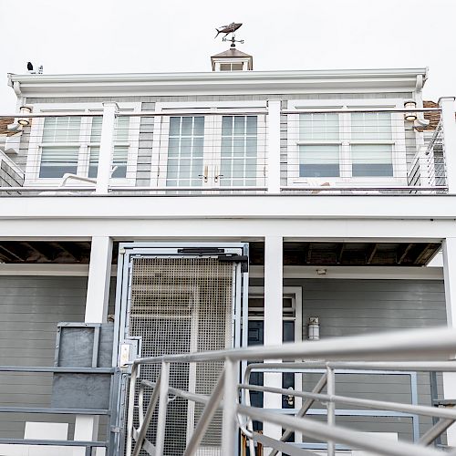 A modern, two-story beach house with large windows and a deck, featuring a weather vane on top and a seagull perched on the roof.