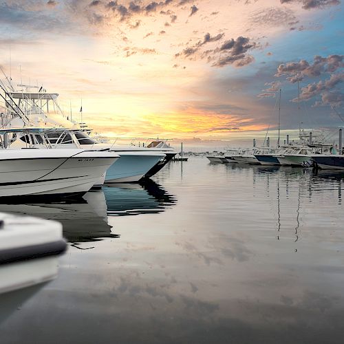 Several boats are docked in a calm marina, illuminated by a picturesque sunset reflecting on the water, with scattered clouds in the sky.