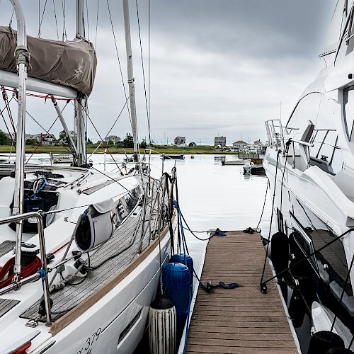 Two boats are docked at a pier near a calm body of water, with a cloudy sky in the background.