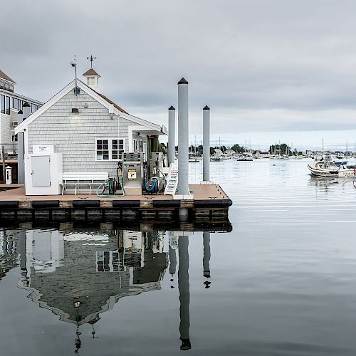 A calm marina with buildings on a dock, boats in the water, and overcast skies reflecting on the water's surface.