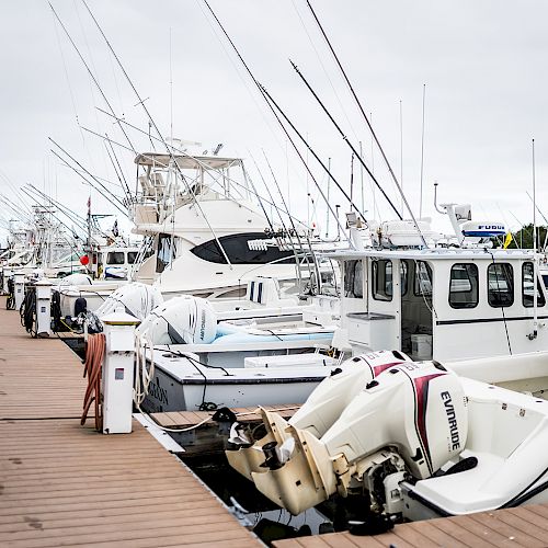 A dock with multiple boats is seen, tied up along the wooden pier on a cloudy day, with fishing rods on some of the boats.