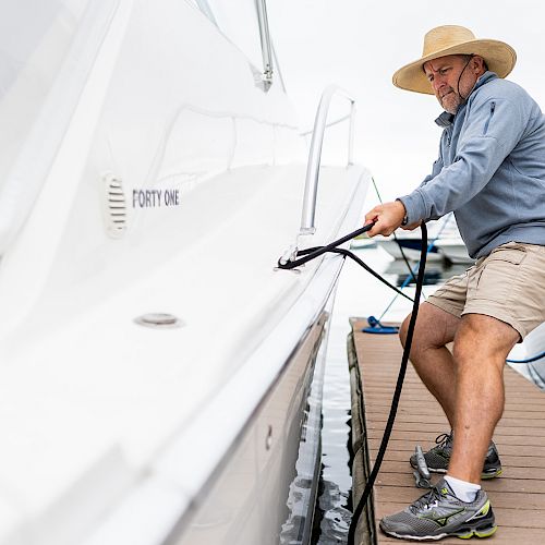 A person wearing a hat and casual clothing is securing a boat to a dock using a rope, with another boat visible in the background.