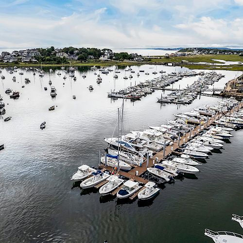 An aerial view of a marina with numerous boats docked, tranquil water, and houses in the background under a partly cloudy sky.