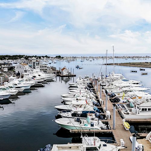 A marina filled with numerous docked boats and yachts, with coastal buildings and a calm sea in the background, under a partly cloudy sky.