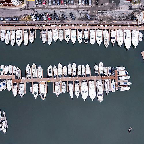 An aerial view of a marina with numerous boats docked at piers in an organized manner, while cars are parked along the waterfront.