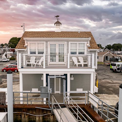 A two-story waterfront building with a small balcony and several chairs, a wooden dock, and parked cars in the background during a colorful sunset.