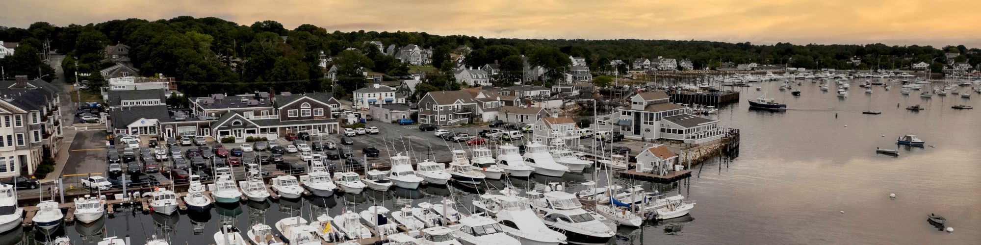 A picturesque marina filled with numerous boats and yachts, set against a serene sunset backdrop with calm waters and a small village in the distance.