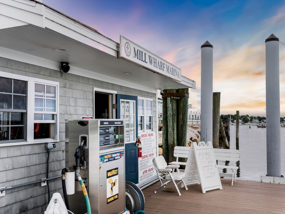 A marina fuel dock at &ldquo;Mill Wharf Marina&rdquo; with fuel pumps, chairs, and boats by the waterfront under a pastel sunset sky.
