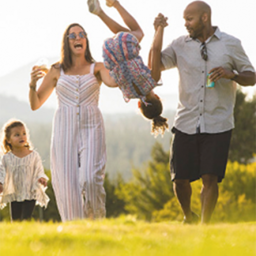 A family, with two adults and two children, are outside in a grassy area. One child is being swung by the adults, while the other walks beside them.