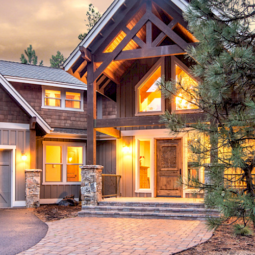 A large modern house with warm interior lighting, two garage doors, and a paved driveway, surrounded by trees during dusk.