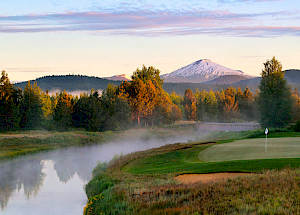 The image shows a golf course with a lush green putting green, a serene river, trees, and a snow-capped mountain in the background under a pastel sky.