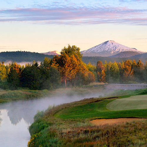 The image shows a golf course with a lush green putting green, a serene river, trees, and a snow-capped mountain in the background under a pastel sky.