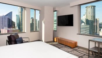 A modern hotel room with large windows, city views, a bed, a TV, an armchair, a wooden table, and a bedside table by the windows.