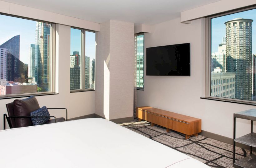 A modern hotel room with large windows, city views, a bed, a TV, an armchair, a wooden table, and a bedside table by the windows.