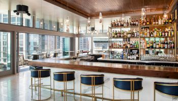 A modern bar with a wooden countertop, stylish barstools, and shelves stocked with various bottles of alcohol. Large windows provide a city view.