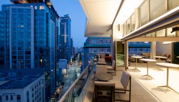 A modern balcony overlooks a city street lined with tall buildings, featuring outdoor seating and tables under warm evening lighting.
