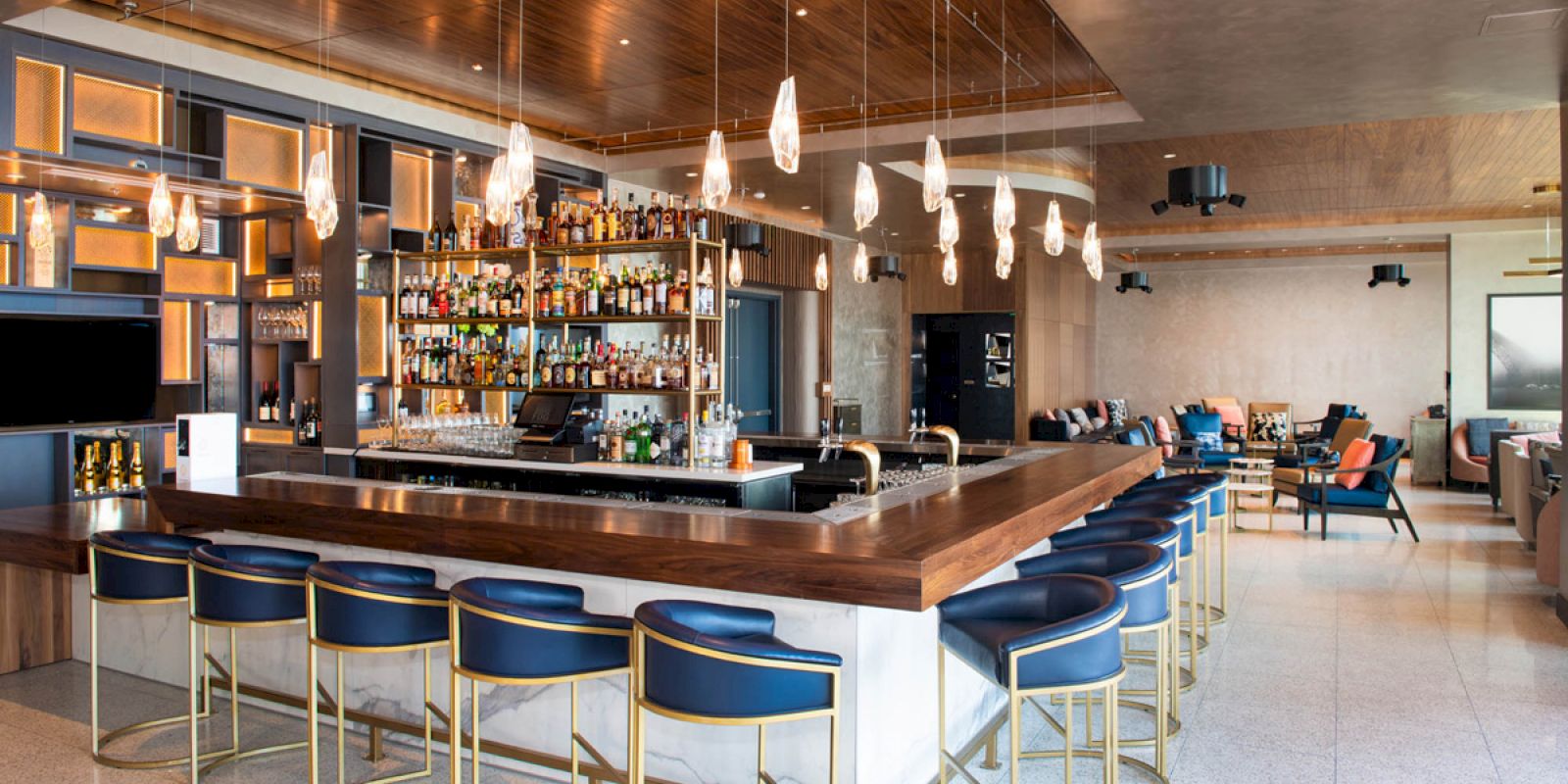 A modern bar with blue barstools, a well-stocked back bar, pendant lights, and a lounge area with seating in the background.