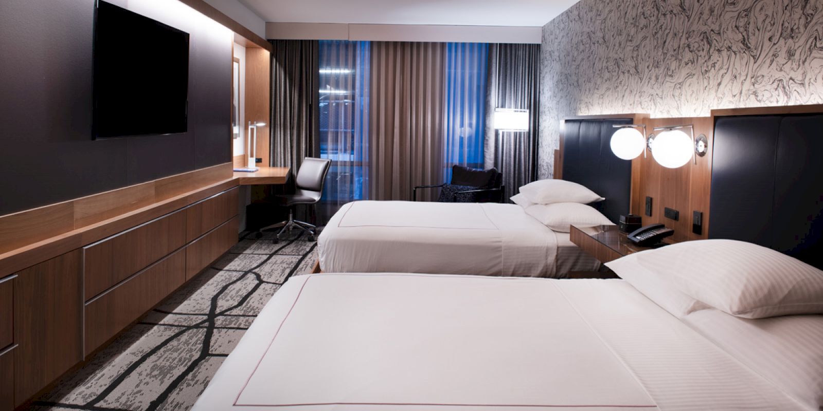 A modern hotel room with two neatly made beds, a wall-mounted TV, a desk with a chair, and stylish lighting.