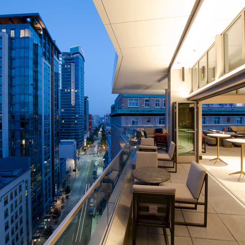 A modern cityscape at dusk with high-rise buildings, viewed from an open, stylish balcony featuring tables, chairs, and soft lighting.