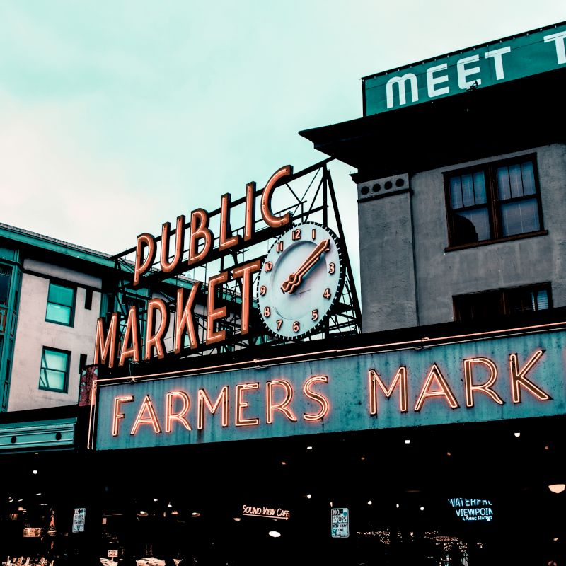 The image shows a busy public market with neon signs saying "PUBLIC MARKET" and "FARMERS MARKET" on a building. The atmosphere seems lively and urban.