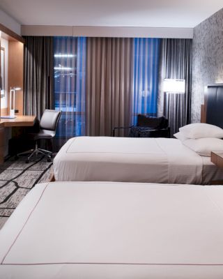 The image shows a modern hotel room with two neatly made beds, a wall-mounted TV, a desk with a chair, and stylish decor with ample lighting.