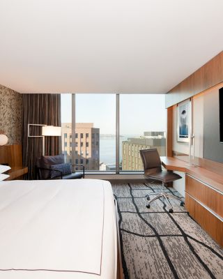 A modern hotel room with a large bed, a wall-mounted TV, a desk with a chair, and a window offering a city view.
