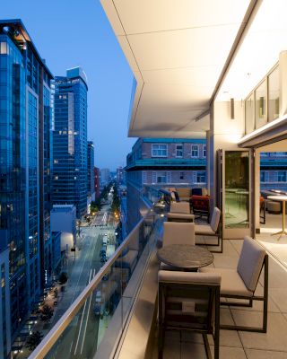 A modern cityscape at dusk with tall buildings and a balcony featuring seating areas and tables, overlooking a bright urban street below.