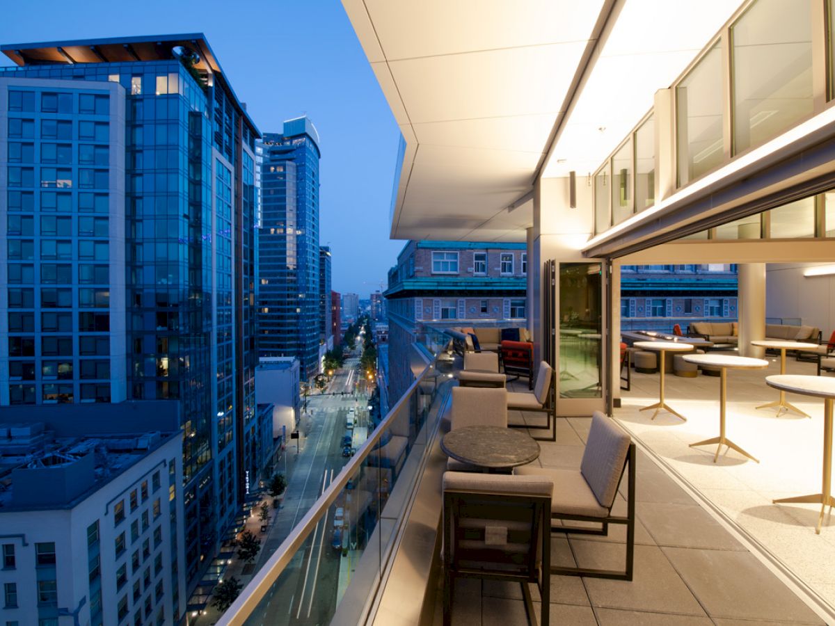 A modern cityscape at dusk with high-rise buildings and a stylish rooftop balcony featuring outdoor seating and tables on the right.