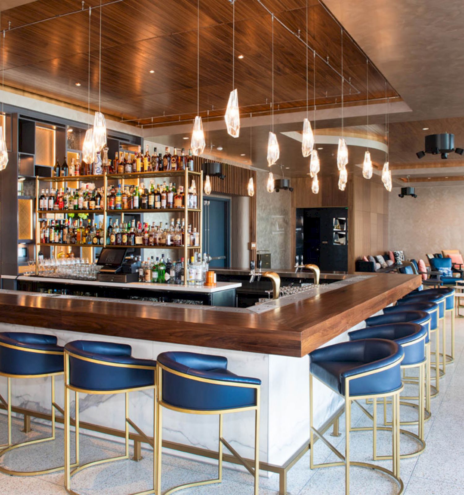 A modern bar with a wooden countertop, blue bar stools, hanging lights, and a well-stocked liquor shelf. There's seating in the background.