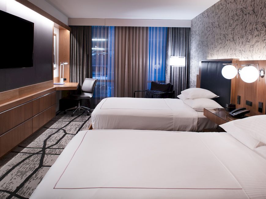 A modern hotel room with two twin beds, a flat-screen TV, a work desk, lamps, and large window drapes. The room has a clean, minimalist design.