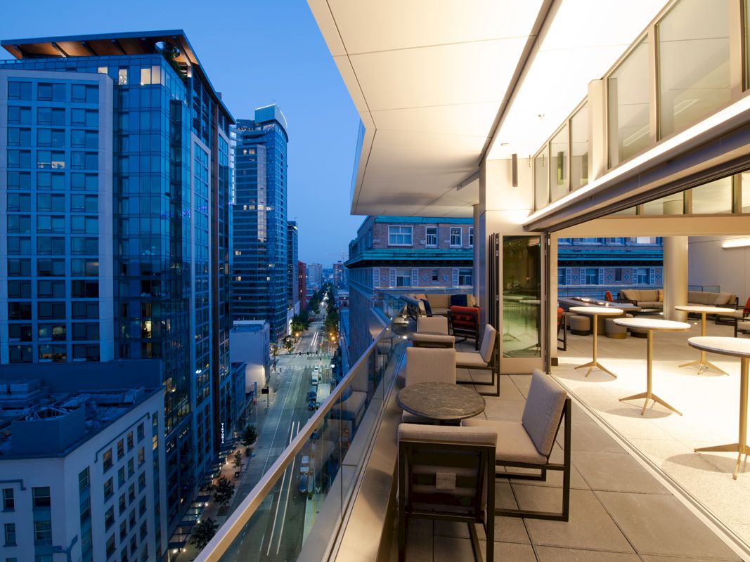 A modern urban balcony at dusk with outdoor seating overlooking a cityscape featuring tall buildings and lit streets, viewed from an elevated perspective.