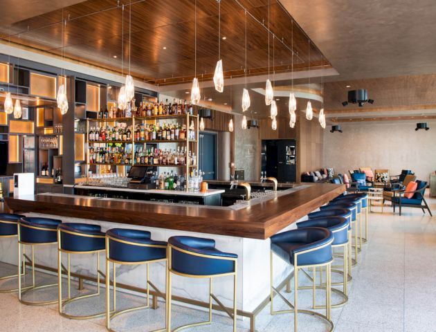 A modern bar with a large assortment of bottles behind the counter, surrounded by barstools and contemporary decor, including hanging lights.
