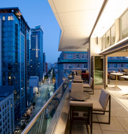 A modern cityscape view from a terrace with tables and chairs, overlooking a street lined with high-rise buildings at dusk.