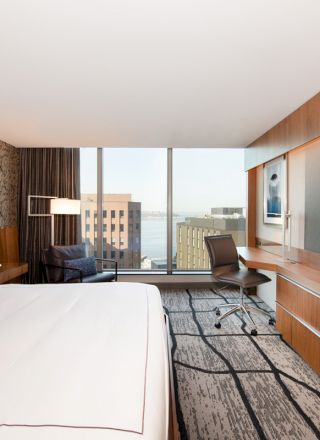 A modern hotel room with a large bed, desk, chair, and TV, featuring a city view through floor-to-ceiling windows, and contemporary decor.