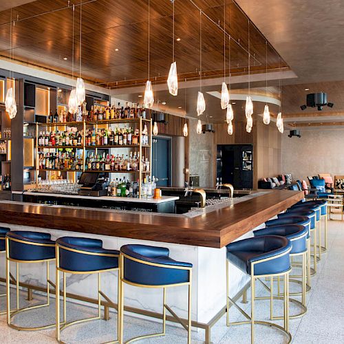 A modern bar with a central island, blue barstools, hanging lights, and shelves filled with various bottles. The interior looks stylish and cozy.