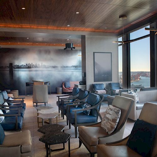 A modern lounge area with plush seating, large windows offering a waterfront view, and a wall mural of a city skyline.