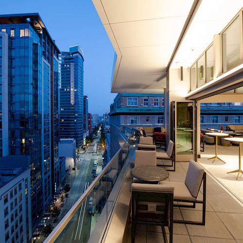 A modern outdoor patio with seating overlooks a cityscape with tall buildings and a street below during the evening.