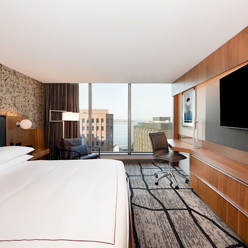 The image shows a modern hotel room with a bed, a desk, a large TV, a chair, and a window overlooking city buildings and water.