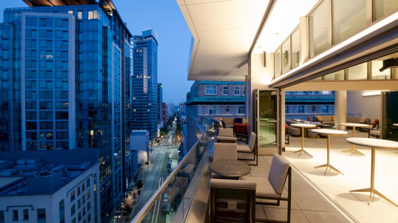 The image shows a modern balcony with tables and chairs overlooking a cityscape with high-rise buildings and a lit-up street at dusk.