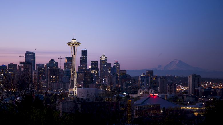 The image shows the Seattle skyline at dusk, with the Space Needle prominently visible and Mount Rainier in the background.