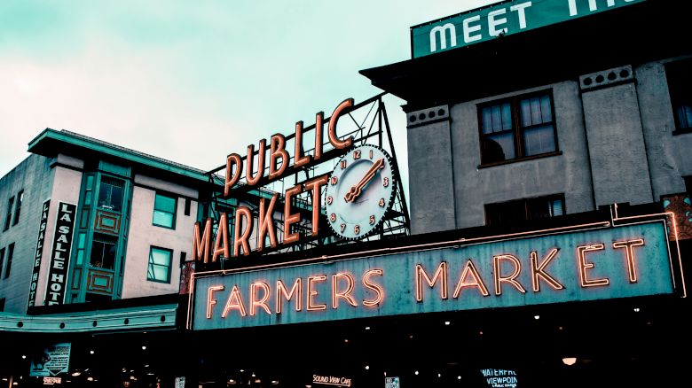 An outdoor sign reads "Public Market" and "Farmers Market" with a clock in the center; nearby buildings display partial signs including a hotel.