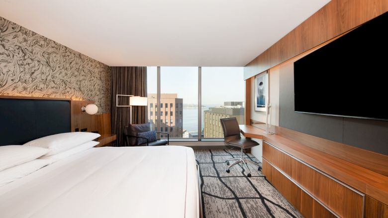 The image shows a modern hotel room with a large bed, a desk, a chair, a flat-screen TV, and a window with a city and water view.