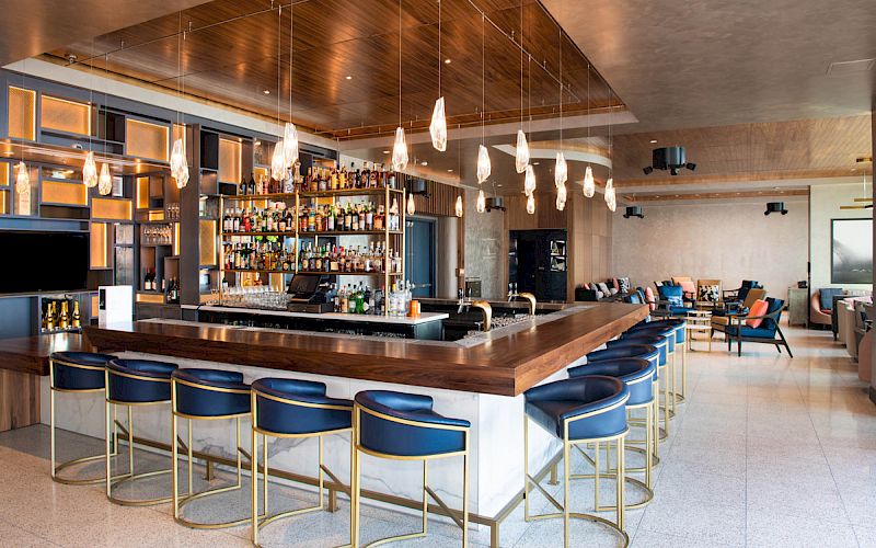 A modern bar with a well-stocked bar counter, stylish overhead lighting, and blue cushioned bar stools, in a chic lounge setting.