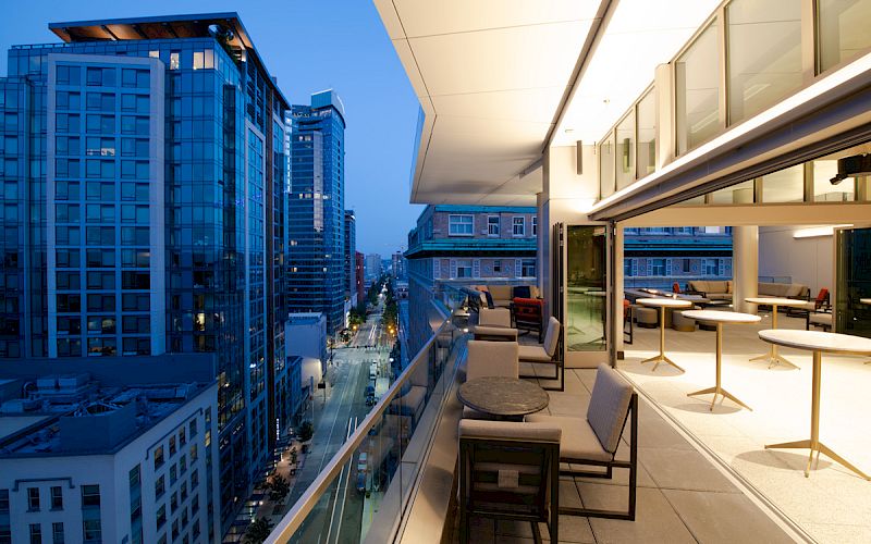 This image shows a modern rooftop terrace with seating and tables, overlooking a cityscape with high-rise buildings at dusk.