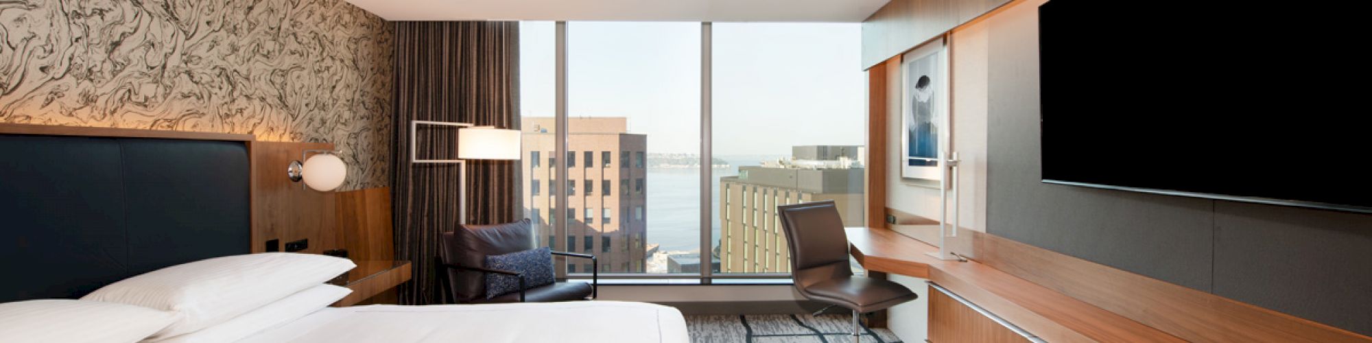 A modern hotel room with a large bed, a desk with a chair, a big TV, and a window showcasing city and water views outside.