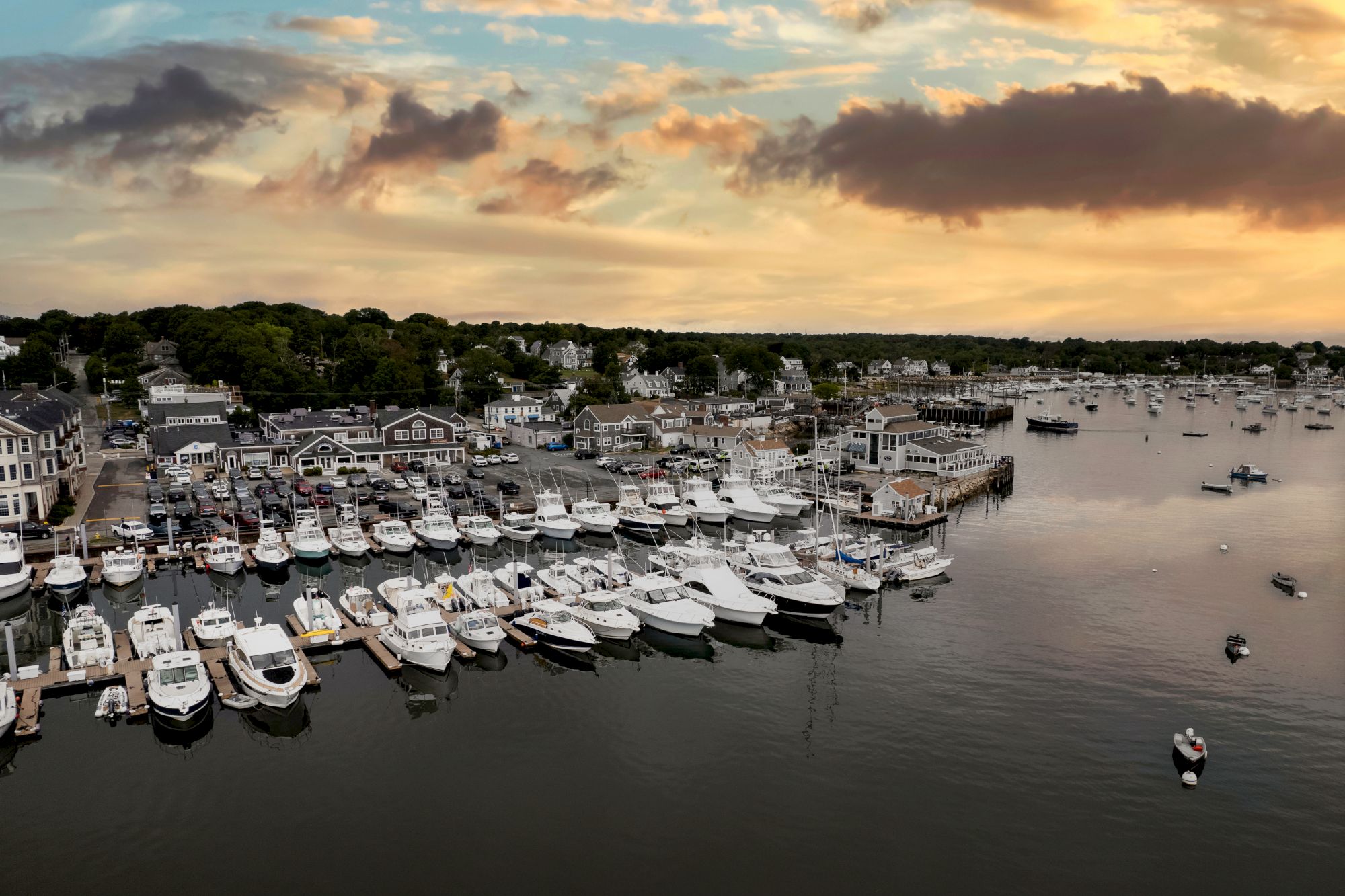 A marina filled with boats and yachts during sunset, with a small town and dense tree line in the background. The water is calm and serene.