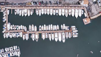 An aerial view of a marina with numerous boats docked along piers, surrounded by calm water. Cars are parked alongside the dock.