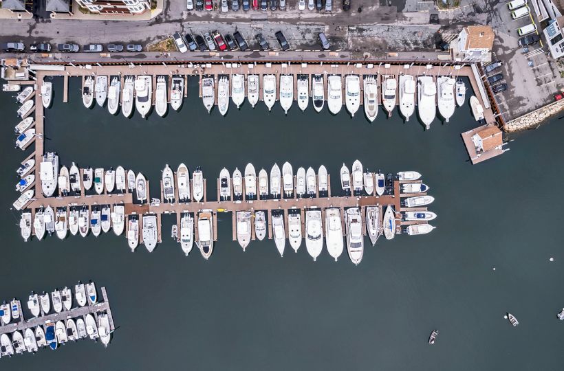 An aerial view of a marina with numerous boats docked along piers, surrounded by calm water. Cars are parked alongside the dock.