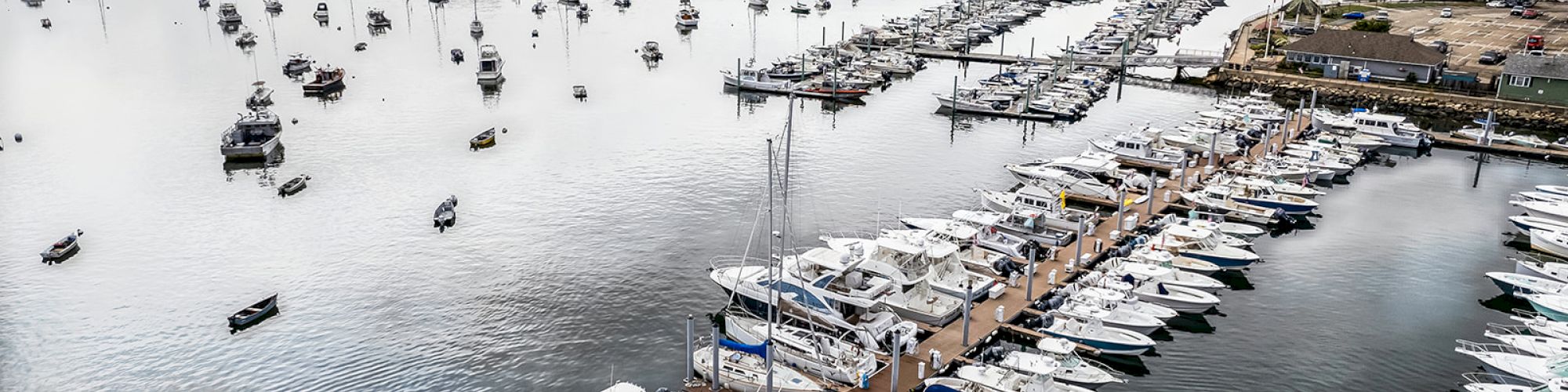 A marina with numerous boats docked, surrounded by calm water and scattered with other boats, reaching towards a distant shoreline under a blue sky.