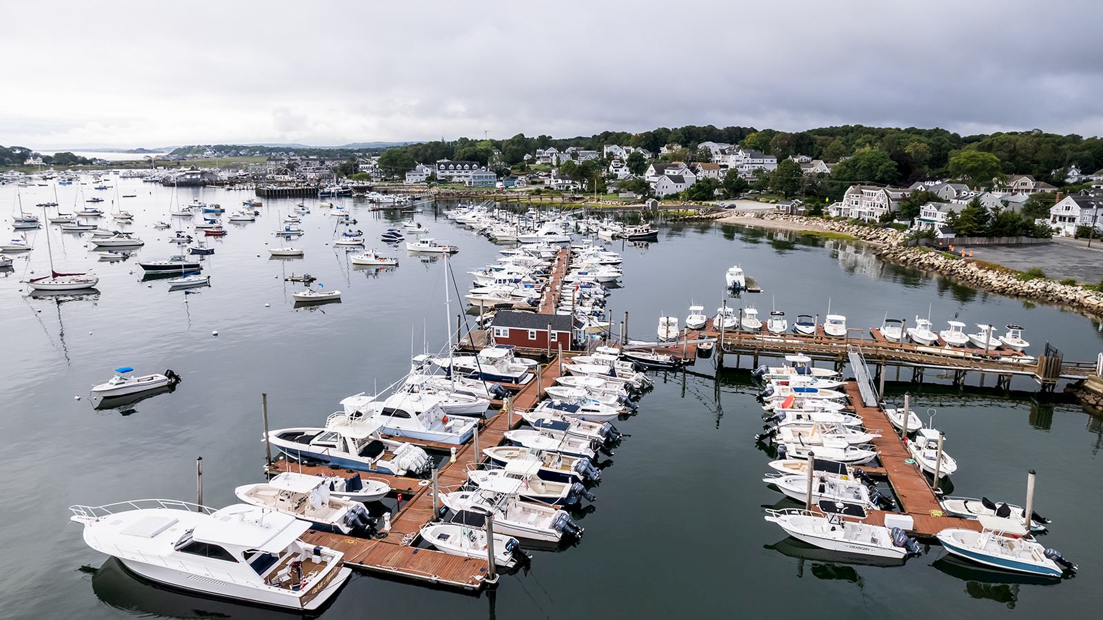 An aerial view of a marina filled with boats docked along wooden piers, with a coastal town and numerous other boats in the background.